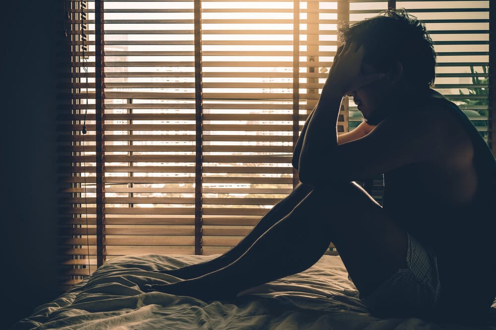 Man sitting on a bed with his head in his hand by a sunlit window
